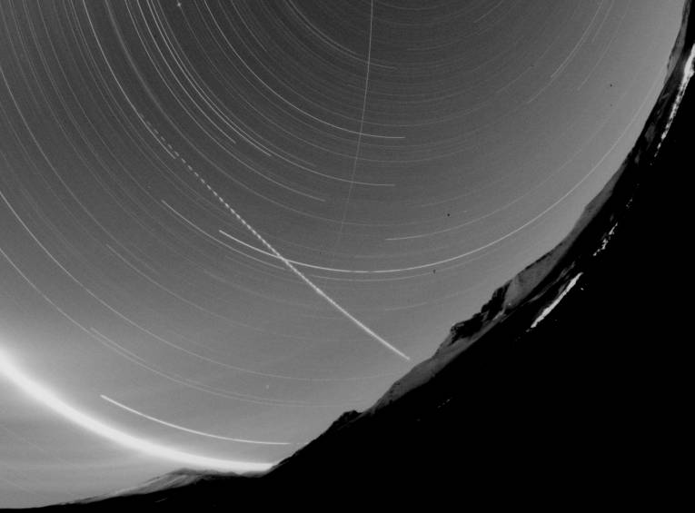 Record from the bolide camera of the Astronomical Institute SAS from the site in Stará Lesná. The picture shows visible circular traces of stars during a couple of hours´ exposition. The clear arch on the left is the trace of the Moon.  The interrupted trace belongs to the meteor (interruptions serve to determine the velocity of the body in the atmosphere), the continuous line from top to bottom is the trace of the artificial Earth satellite. Sidelong on the right the massif of the Lomnický Štít can be seen